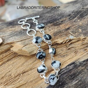 Könnte beinhalten: Ein silbernes Armband mit ovalen, schwarz-weiß gesprenkelten Steinen. Das Armband ist durch silberne Glieder verbunden und hat einen Knebelverschluss. Es wird auf einer Holzoberfläche präsentiert.