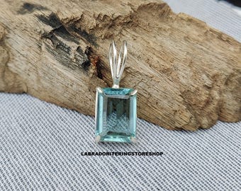 May include: A rectangular, pale blue gemstone pendant with a silver bail. The pendant is set in a silver frame and is displayed on a piece of driftwood. The text "LABRADORITERINGSTORESHOP" is visible at the bottom.