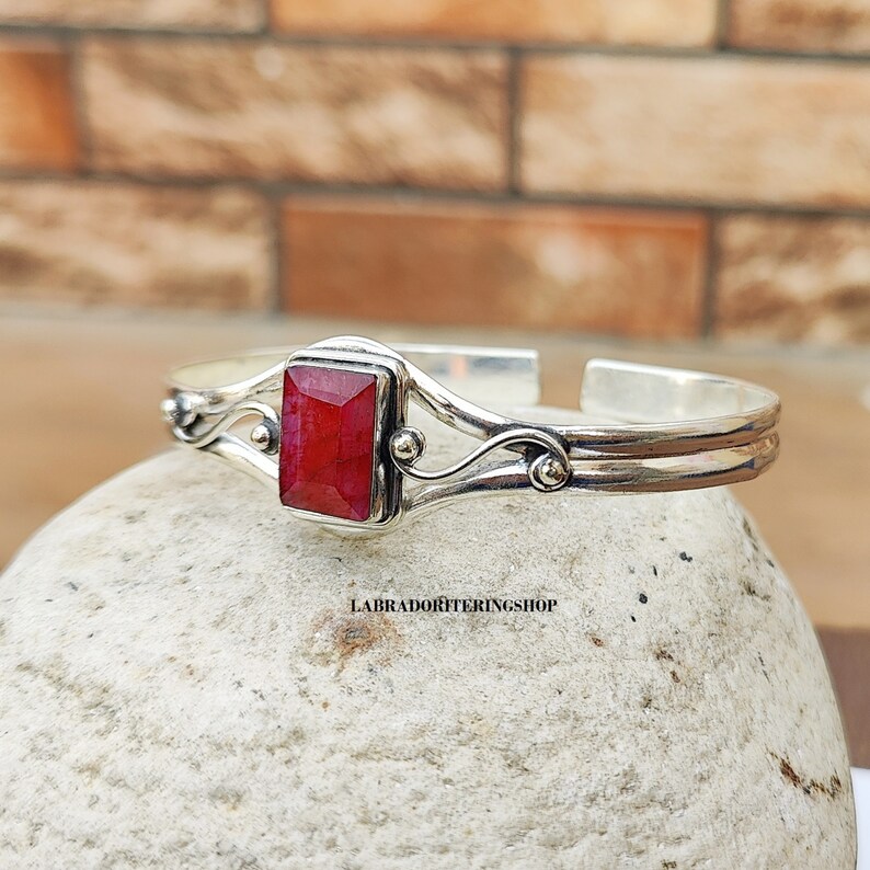 May include: A silver bangle bracelet with a red gemstone set in a silver bezel. The bracelet has a delicate, ornate design with swirls and small beads.
