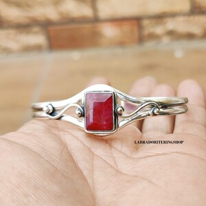 May include: A silver bangle bracelet with a red gemstone set in a rectangular shape. The bracelet has a delicate, flowing design with small beads on each side of the gemstone.