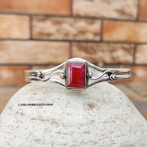 May include: A silver bangle bracelet with a red gemstone set in a silver bezel. The bracelet has a delicate, ornate design with swirls and beads.