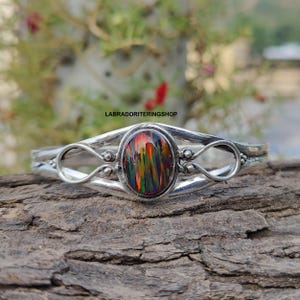 Può includere: Un bracciale in argento con una gemma ovale multicolore. La gemma mostra una vibrante gamma di colori tra cui rosso, verde e arancione. Il bracciale ha un design intricato con dettagli ad anello e il testo "LABRADORITERINGSHOP".