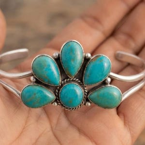 May include: A silver cuff bracelet featuring five teardrop-shaped turquoise stones and one central round turquoise stone. The stones are set in a fan-like arrangement with a twisted silver wire border surrounding the centre stone.