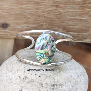 Puede incluir: Pulsera de plata con una gran incrustación ovalada de concha de abulón. La concha tiene colores iridiscentes verdes, azules y morados.