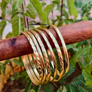 May include: A set of six gold-colored bangles with a hammered texture, displayed on a wooden dowel against a backdrop of green foliage. The bangles have a shiny finish and a simple, elegant design. The text "LABRADORITERINGSHOP" is visible.