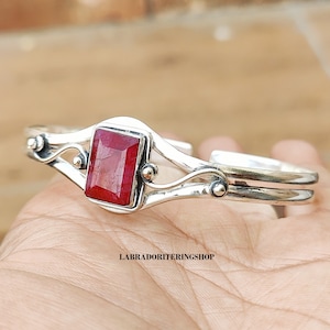 May include: Silver cuff bracelet with a red gemstone set in a silver bezel. The bracelet has a delicate, ornate design with two curved bands and small silver beads.