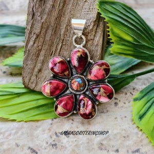 May include: A silver pendant with a flower design. The pendant features red, pink, and gold-coloured stones arranged in a floral pattern. The centre stone is a mix of blue and brown. The text "LABRADORITERINGSHOP" is visible at the bottom.