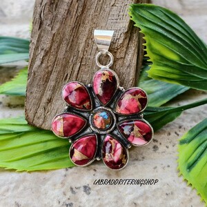 May include: A silver pendant in the shape of a flower, featuring red, pink, and gold-coloured stones. The central stone is a mix of blue, pink, and gold. The pendant is displayed on a wooden surface. The text "LABRADORITERINGSHOP" is visible.