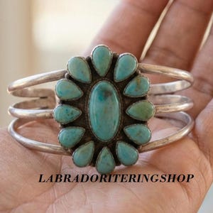 Könnte beinhalten: Ein Silberarmreif mit einem zentralen ovalen Türkisstein, der von zwölf kleineren, tropfenförmigen Türkissteinen in einem Blumenmuster umgeben ist. Das Armband besteht aus drei parallelen Silberdrähten, die an den Seiten zusammenlaufen.