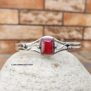May include: A silver bangle bracelet with a red gemstone set in a square shape. The bracelet has a delicate, ornate design with swirls and beads.