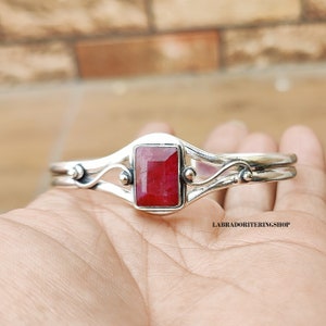 May include: A silver bracelet with a red gemstone set in a rectangular shape. The bracelet has a delicate, curved design with small accents.