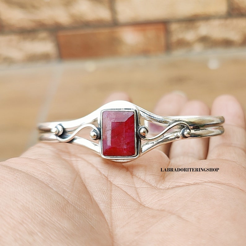 May include: A silver bracelet with a red gemstone set in a rectangular shape. The bracelet has a delicate, curved design with small accents.