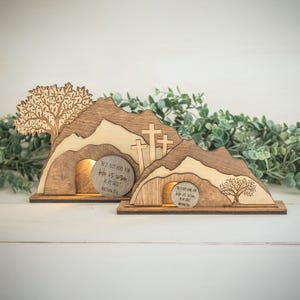 May include: Wooden Easter scene featuring a mountain with a tomb and three crosses. The tomb has a round stone with the text "He is not here for He is risen, as He said." A tree is on the left side. The scene is made of light and dark brown wood.