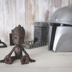 Peut inclure: Une figurine de Groot brune et verte assise sur une surface blanche. La figurine porte une tenue brune et verte et a un visage brun. Il y a un casque argenté et une structure en pierre grise en arrière-plan.