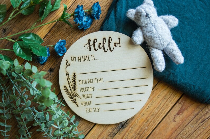 May include: A round wooden sign with the text "Hello! My name is..." and blank lines for birth date/time, location, height, weight, and head size. The sign is decorated with a floral design.