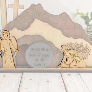 May include: Wooden Easter scene featuring an angel, crosses, and figures near a tomb. The background depicts a mountain range. The text on the tomb reads "HE IS NOT HERE FOR He is risen, AS HE SAID MATTHEW 28:6."