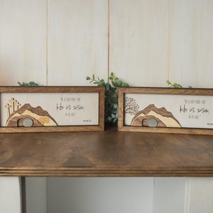 May include: Two wooden framed art pieces with a religious theme. Each features a mountain scene with a tomb and the text "He is not here for He is risen, as He said." The frames are brown, and the background is a light beige.