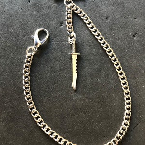 Puede incluir: Una cadena de reloj de bolsillo de plata con un dije de cuchillo. La cadena está hecha de un estilo de eslabón grueso y de tipo curb. El dije de cuchillo es un diseño simple y plano.