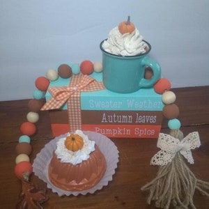 May include: A teal mug with whipped cream and a small pumpkin on top, a brown and orange cake with whipped cream and a small pumpkin on top, and a wooden box with the words "Sweater Weather", "Autumn Leaves", and "Pumpkin Spice" on it. The box is decorated with a white and red gingham ribbon and a beaded garland with orange, brown, and teal beads.