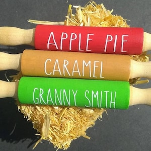 May include: Three wooden rolling pins with different colored handles and painted text. The top rolling pin is red with the text "APPLE PIE", the middle rolling pin is brown with the text "CARAMEL", and the bottom rolling pin is green with the text "GRANNY SMITH".
