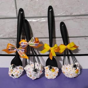 May include: Four Halloween-themed kitchen whisks. Each whisk has a black handle and is decorated with white frosting, candy sprinkles, and a black spider. Orange and yellow ribbons with "BOO!" text adorn the handles.