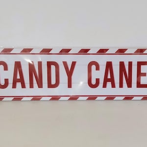 May include: A rectangular sign with a white background and red and white striped border. The sign reads "CANDY CANE LN" in red block letters. Candy cane and bow illustrations are on the sign.