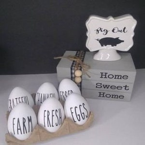 May include: A gray wooden box with the words "Home Sweet Home" painted on it. The box is decorated with a black and white checkered ribbon and wooden beads. A white ceramic sign with a pig and the words "Pig Out" is on top of the box. Six white eggs with different words written on them are in a brown egg carton.