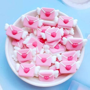 May include: A bowl filled with pink and white resin charms shaped like envelopes with hearts and wings. The charms are perfect for adding a touch of whimsy to crafts and jewelry.