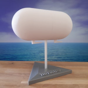 Könnte beinhalten: Ein weißes, kapselartiges Objekt ist auf einem weißen Ständer montiert, der an einer grauen, dreieckigen Basis befestigt ist. Auf der Basis steht der Text "Tic-Tac-Up". Der Hintergrund zeigt eine Meereslandschaft.
