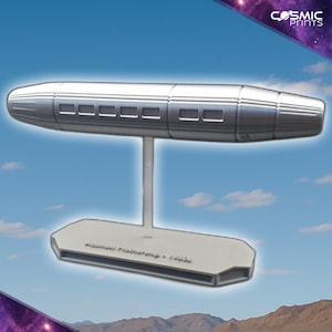 Puede incluir: Modelo plateado de una nave espacial, sostenido por un soporte vertical delgado y una base hexagonal blanca. La nave espacial tiene un cuerpo cilíndrico con ventanas rectangulares. La base tiene el texto "Alien Mothership - 1950s".