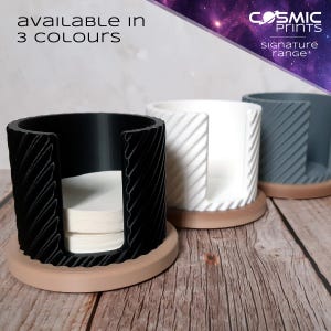 Può includere: Tre supporti cilindrici in nero, bianco e grigio, ciascuno con un design strutturato e una base beige. I supporti sono progettati per contenere filtri di carta rotondi. Il testo "AVAILABLE in 3 COLOURS" è visibile.
