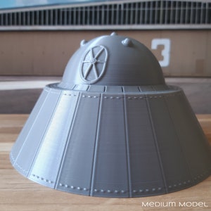 Puede incluir: Modelo gris impreso en 3D de un platillo volante. El modelo tiene una parte superior redondeada con una ventana circular y varias pequeñas protuberancias. La base es acampanada y tiene paneles verticales con remaches simulados. El texto "MEDIUM MODEL SHOWN" está en la parte inferior.