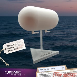 May include: A white, capsule-shaped object on a display stand. The stand has a triangular base with the text "Tic Tac UAP" and a tag that says "Display Stand Included". The background is a seascape.