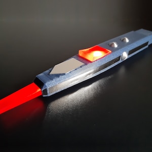 May include: A futuristic, blue and gray device with a glowing red rectangular tip. The device has a silver accent, two silver buttons, and a red, diamond-shaped light. The overall design suggests a science fiction theme.