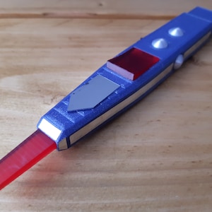 May include: A blue and silver device with a red rectangular tip and a silver arrow-shaped detail. The device has two silver buttons and a red square. The object is on a wooden surface.