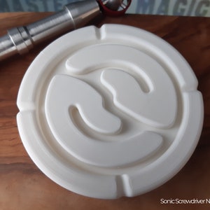 Può includere: Un oggetto circolare bianco con un design stilizzato in rilievo. L'oggetto presenta una serie di anelli concentrici e un motivo centrale curvo. Un oggetto metallico color argento è parzialmente visibile sullo sfondo. Il testo "Sonic Screwdriver Not Included" è in basso.