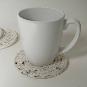 Pu&ograve; includere: Una tazza di ceramica bianca con manico su un sottobicchiere all'uncinetto. Il sottobicchiere &egrave; fatto di filato beige con macchie nere e marroni.