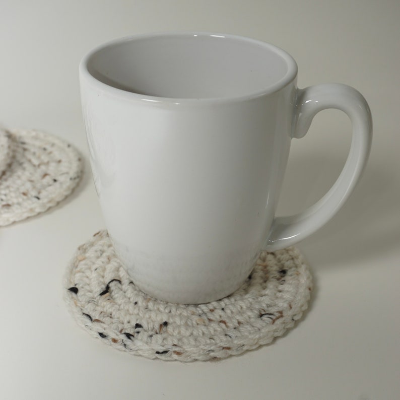 Pu&ograve; includere: Una tazza di ceramica bianca con manico su un sottobicchiere all'uncinetto. Il sottobicchiere &egrave; fatto di filato beige con macchie nere e marroni.