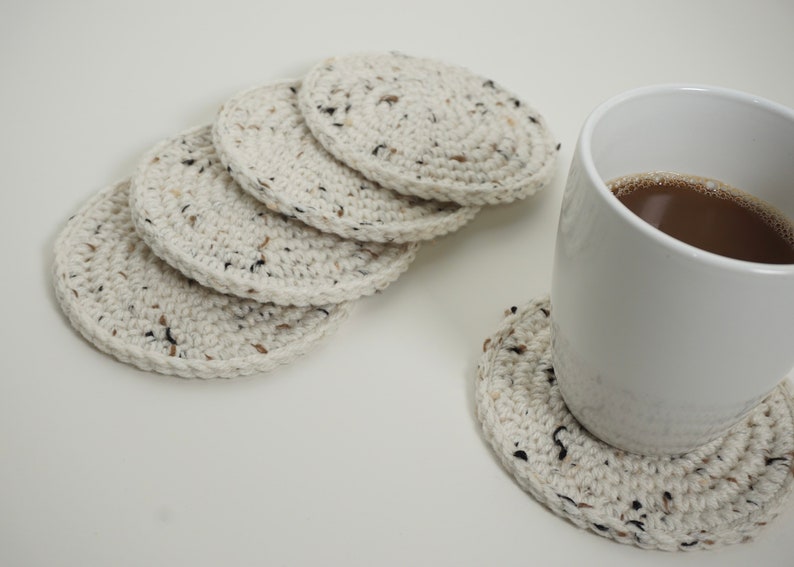 Pu&ograve; includere: Cinque sottobicchieri in crochet bianco con macchie nere e marroni, impilati su una superficie bianca. Una tazza in ceramica bianca con caff&egrave; marrone si trova su un sesto sottobicchiere.