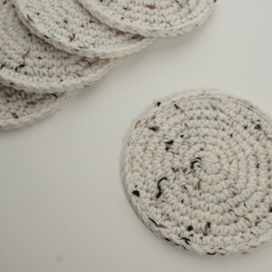 Pu&ograve; includere: Un set di sei sottobicchieri in crochet bianco con macchie marroni e nere. I sottobicchieri sono rotondi e hanno una superficie testurizzata.