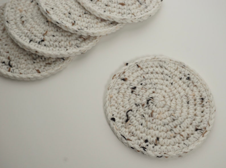 Pu&ograve; includere: Un set di sei sottobicchieri in crochet bianco con macchie marroni e nere. I sottobicchieri sono rotondi e hanno una superficie testurizzata.