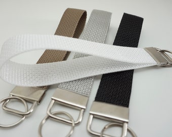 May include: A collection of keychains in various colors: white, tan, silver, and black. Each keychain features a woven fabric strap attached to a silver-colored metal ring and a rectangular metal piece.