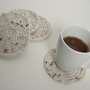 Pu&ograve; includere: Una tazza da caff&egrave; in ceramica bianca con un liquido marrone al suo interno, appoggiata su un sottobicchiere all'uncinetto. Ci sono altri tre sottobicchieri all'uncinetto sullo sfondo.