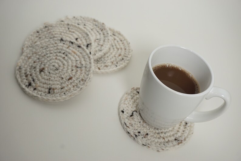 Pu&ograve; includere: Una tazza da caff&egrave; in ceramica bianca con un liquido marrone al suo interno, appoggiata su un sottobicchiere all'uncinetto. Ci sono altri tre sottobicchieri all'uncinetto sullo sfondo.