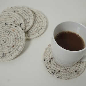Pu&ograve; includere: Quattro sottobicchieri all'uncinetto di colore crema con macchie nere, uno dei quali &egrave; utilizzato sotto una tazza bianca piena di caff&egrave; marrone.