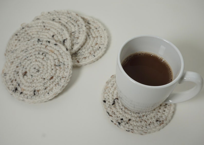 Pu&ograve; includere: Quattro sottobicchieri all'uncinetto di colore crema con macchie nere, uno dei quali &egrave; utilizzato sotto una tazza bianca piena di caff&egrave; marrone.