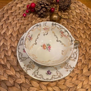 Könnte beinhalten: Ein cremefarbenes Tee-Service mit Tasse und Untertasse, verziert mit goldenen Verzierungen und floralen Mustern in Rosa, Lila und Gelb. Das Set steht auf einem geflochtenen Platzset, dekoriert mit Tannenzapfen und roten Beeren.