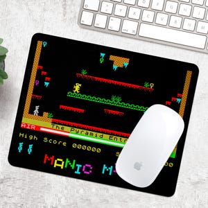 May include: A black and yellow mouse mat with a retro video game design. The game is called "Manic Miner" and features a pyramid, a character, and a high score. The mouse mat is designed to look like an old-school arcade game.