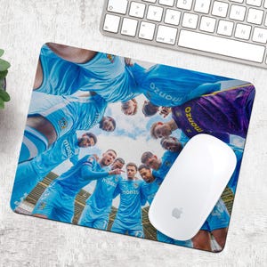 May include: A rectangular mouse mat featuring a team of soccer players in light blue uniforms, huddled together. The word "monzo" is visible on the uniforms. A white computer mouse is on the right side of the mat.