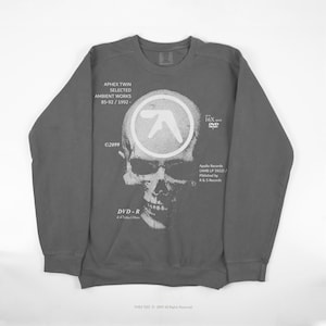 May include: Gray sweatshirt featuring a white skull graphic with the Aphex Twin logo and text. The text includes "APHEX TWIN SELECTED AMBIENT WORKS 85-92/1992-" and "DVD-R". The sweatshirt has a crew neck and long sleeves.